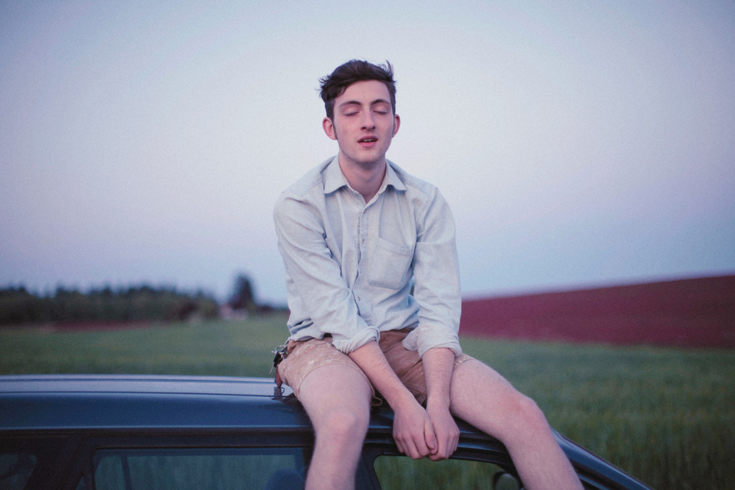 a young man wearing a button up and shorts sitting on the roof of a car