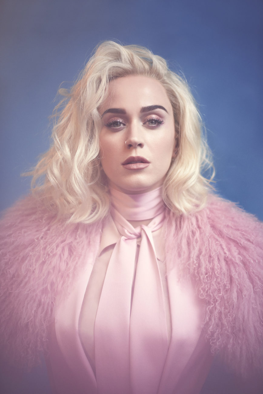a headshot of katy perry featuring blond hair, a pink suit, and a pink fuzzy jacket against a blue background