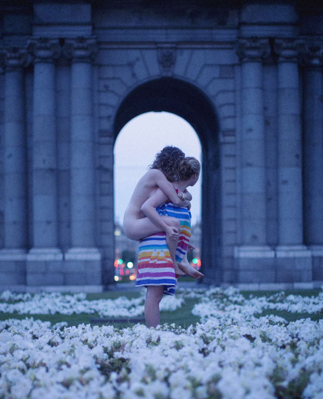 a woman in a dress carrying a naked woman on her back in a field of flowers with a large stone arch behind them